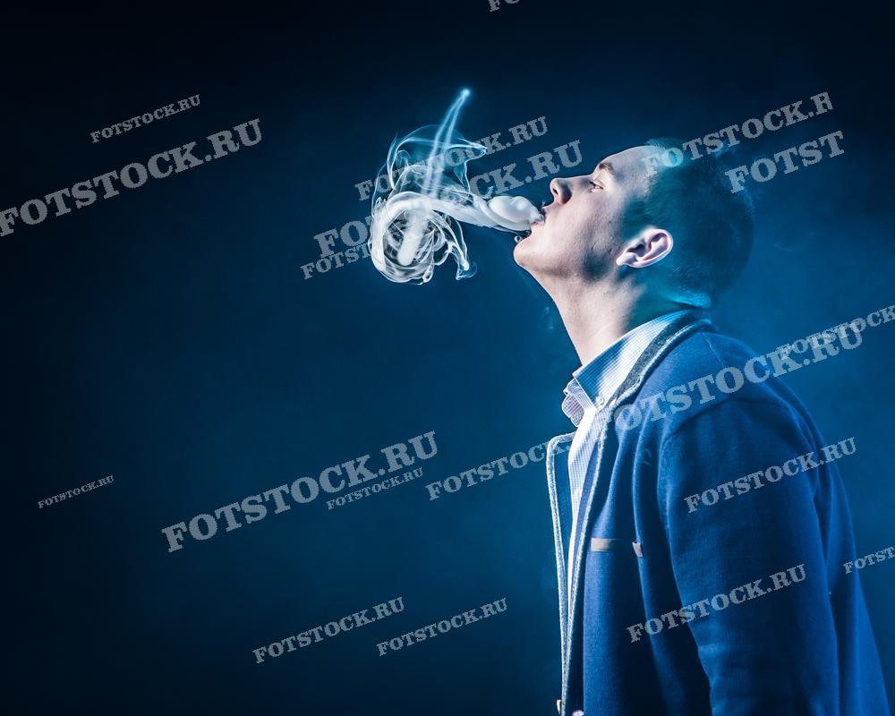 Молодой человек в темной одежде выдыхает клубы дыма, создавая эффектный визуальный образ на фоне тёмного пространства. Фотография иллюстрирует процесс вейпинга и создаёт атмосферу современного стиля жизни.